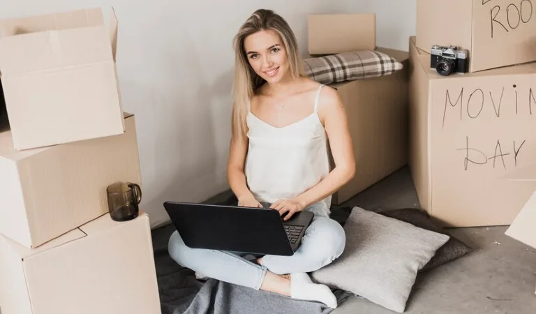 Lady is working on her laptop sitting on floor with moving boxes around her.